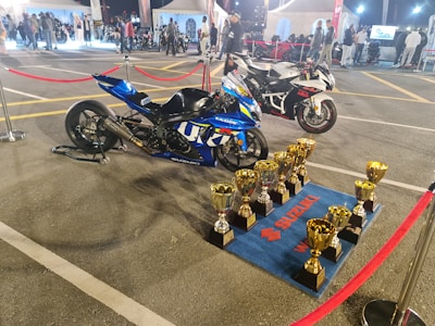 Two high-performance motorcycles are displayed on a parking lot surrounded by red rope barriers. In front of the blue motorcycle, several golden trophies are neatly arranged on a blue mat branded with Suzuki logos. The background features tents, people, and additional motorcycles, suggesting a competitive or exhibition event.