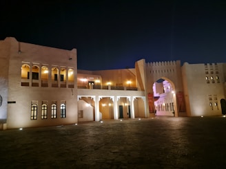 An architectural structure featuring traditional Middle Eastern design elements, including arched windows and a large archway. The building is illuminated with soft lights that cast warm tones against the stone facade. The scene is set at night, creating a serene and calm environment.