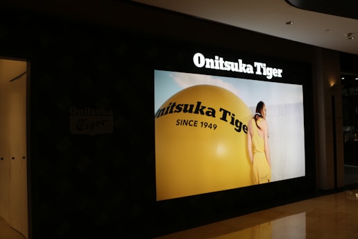 A store display showcases a large advertisement for Onitsuka Tiger, featuring a person in a yellow outfit standing next to a giant yellow sphere with the brand's name and 'Since 1949' written on it.
