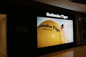 A store display showcases a large advertisement for Onitsuka Tiger, featuring a person in a yellow outfit standing next to a giant yellow sphere with the brand's name and 'Since 1949' written on it.