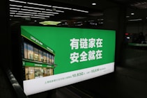 A large illuminated advertisement is displayed in an indoor setting, primarily showing a real estate company's branding. The sign features a green background with white text and includes an image of a building with the company's name, Lianjia, on it.