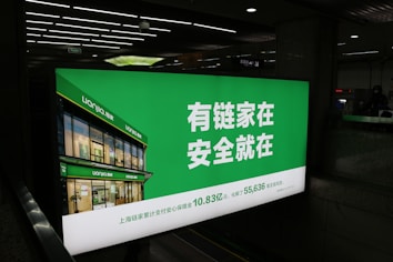 A large illuminated advertisement is displayed in an indoor setting, primarily showing a real estate company's branding. The sign features a green background with white text and includes an image of a building with the company's name, Lianjia, on it.