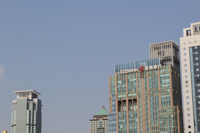 Several modern skyscrapers with various architectural designs rise against a clear blue sky. The buildings feature a mix of glass and metal facades, with one displaying the logo of a bank.