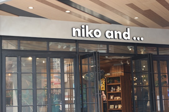 The entrance of a store featuring tall black-framed glass doors and a sign reading 'niko and ...'. Inside, wooden shelves display various items, and warm lighting illuminates the space. The ceiling is designed with wooden panels.