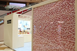 A modern retail display area in a shopping mall features a large wall filled with an intricate pattern of pink roses. Adjacent to the floral display is a beige booth with minimalist design, showcasing a range of products. The interior of the booth has shelves with various items and a lighted background with the image of a person in a blue shirt.