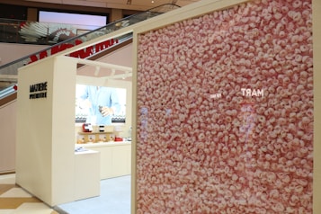 A modern retail display area in a shopping mall features a large wall filled with an intricate pattern of pink roses. Adjacent to the floral display is a beige booth with minimalist design, showcasing a range of products. The interior of the booth has shelves with various items and a lighted background with the image of a person in a blue shirt.