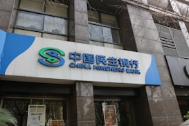 The image features the exterior of a bank building, specifically the China Minsheng Bank. The signage is prominently displayed with both Chinese characters and English lettering, all in a modern, stylized font. The facade is constructed with glass and dark stone panels, and there's a partial view of tree branches overhead.