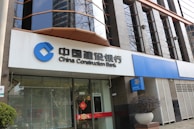 A modern bank building with a prominent sign in both Chinese and English for China Construction Bank. The facade features large, reflective windows and a clean, structured architectural design. Potted plants are placed near the entrance, giving it an inviting look.