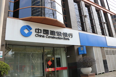 A modern bank building with a prominent sign in both Chinese and English for China Construction Bank. The facade features large, reflective windows and a clean, structured architectural design. Potted plants are placed near the entrance, giving it an inviting look.