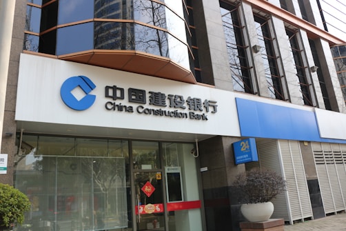 A modern bank building with a prominent sign in both Chinese and English for China Construction Bank. The facade features large, reflective windows and a clean, structured architectural design. Potted plants are placed near the entrance, giving it an inviting look.