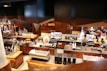 A modern coffee shop with a warm, wooden interior. Several baristas are behind the counter, preparing coffee and other beverages. The counters are equipped with various coffee-making machines and supplies. In the background, a few customers are seated, enjoying their drinks. The shop has a cozy and inviting atmosphere with a mix of contemporary and rustic design elements.