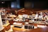 A modern coffee shop with a warm, wooden interior. Several baristas are behind the counter, preparing coffee and other beverages. The counters are equipped with various coffee-making machines and supplies. In the background, a few customers are seated, enjoying their drinks. The shop has a cozy and inviting atmosphere with a mix of contemporary and rustic design elements.