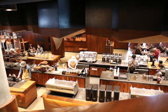 A modern coffee shop with a warm, wooden interior. Several baristas are behind the counter, preparing coffee and other beverages. The counters are equipped with various coffee-making machines and supplies. In the background, a few customers are seated, enjoying their drinks. The shop has a cozy and inviting atmosphere with a mix of contemporary and rustic design elements.