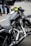 A close-up view of a sleek, black motorcycle with chrome detailing and a leather seat. The handlebars have an attached rear-view mirror and a bright yellow cloth is wrapped around one side. In the background, people are wearing jeans and leather jackets, standing on a paved surface.