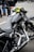 A close-up view of a sleek, black motorcycle with chrome detailing and a leather seat. The handlebars have an attached rear-view mirror and a bright yellow cloth is wrapped around one side. In the background, people are wearing jeans and leather jackets, standing on a paved surface.