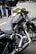 A close-up view of a sleek, black motorcycle with chrome detailing and a leather seat. The handlebars have an attached rear-view mirror and a bright yellow cloth is wrapped around one side. In the background, people are wearing jeans and leather jackets, standing on a paved surface.
