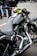 A close-up view of a sleek, black motorcycle with chrome detailing and a leather seat. The handlebars have an attached rear-view mirror and a bright yellow cloth is wrapped around one side. In the background, people are wearing jeans and leather jackets, standing on a paved surface.
