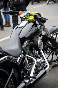 A close-up view of a sleek, black motorcycle with chrome detailing and a leather seat. The handlebars have an attached rear-view mirror and a bright yellow cloth is wrapped around one side. In the background, people are wearing jeans and leather jackets, standing on a paved surface.