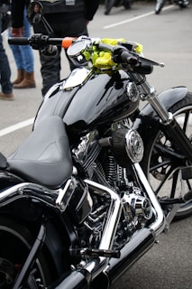 A close-up view of a sleek, black motorcycle with chrome detailing and a leather seat. The handlebars have an attached rear-view mirror and a bright yellow cloth is wrapped around one side. In the background, people are wearing jeans and leather jackets, standing on a paved surface.