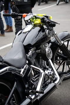 A close-up view of a sleek, black motorcycle with chrome detailing and a leather seat. The handlebars have an attached rear-view mirror and a bright yellow cloth is wrapped around one side. In the background, people are wearing jeans and leather jackets, standing on a paved surface.