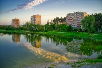 Three mid-rise apartment buildings stand near a serene lake, their reflections mirrored on its calm surface. Lush green trees and grass line the shore, and the sky is partly cloudy with a warm, golden hue from the setting sun.