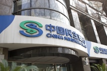 The image features the exterior of a bank building with large signs displaying the name 'China Minsheng Bank' in both Chinese characters and in English. The design includes a logo with the initials 'S' in green and blue. The building's facade consists of reflective glass and concrete elements, with some greenery visible at the bottom.