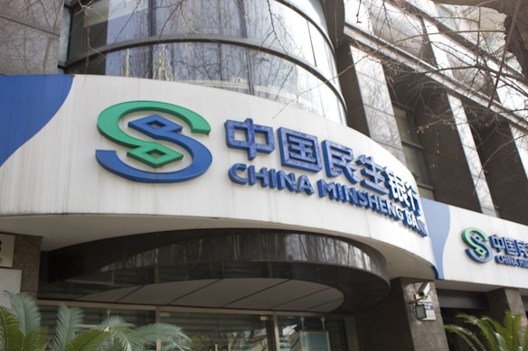 The image features the exterior of a bank building with large signs displaying the name 'China Minsheng Bank' in both Chinese characters and in English. The design includes a logo with the initials 'S' in green and blue. The building's facade consists of reflective glass and concrete elements, with some greenery visible at the bottom.
