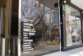 Glass storefront featuring a list of wine varieties in white text. The reflection in the glass shows a street scene with trees, a building, and several yellow rental bicycles. Inside the store, there are tall, silver metal objects, possibly heaters or decor.