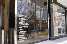 Glass storefront featuring a list of wine varieties in white text. The reflection in the glass shows a street scene with trees, a building, and several yellow rental bicycles. Inside the store, there are tall, silver metal objects, possibly heaters or decor.