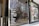 Glass storefront featuring a list of wine varieties in white text. The reflection in the glass shows a street scene with trees, a building, and several yellow rental bicycles. Inside the store, there are tall, silver metal objects, possibly heaters or decor.