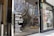 Glass storefront featuring a list of wine varieties in white text. The reflection in the glass shows a street scene with trees, a building, and several yellow rental bicycles. Inside the store, there are tall, silver metal objects, possibly heaters or decor.