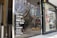 Glass storefront featuring a list of wine varieties in white text. The reflection in the glass shows a street scene with trees, a building, and several yellow rental bicycles. Inside the store, there are tall, silver metal objects, possibly heaters or decor.