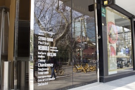 Glass storefront featuring a list of wine varieties in white text. The reflection in the glass shows a street scene with trees, a building, and several yellow rental bicycles. Inside the store, there are tall, silver metal objects, possibly heaters or decor.