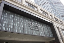 Entrance of HKRI Taikoo Hui, featuring modern architecture with a geometric design. The facade is composed of glass and metal, with visible text in both Chinese and English. A tall building with numerous windows is visible in the background.