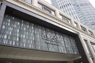 Entrance of HKRI Taikoo Hui, featuring modern architecture with a geometric design. The facade is composed of glass and metal, with visible text in both Chinese and English. A tall building with numerous windows is visible in the background.