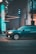 A dark teal SUV is in motion on a city street at night. The background is blurred, suggesting speed, with bright teal and orange lights reflecting off the vehicle. The road appears empty, with an urban environment featuring buildings and streetlights.