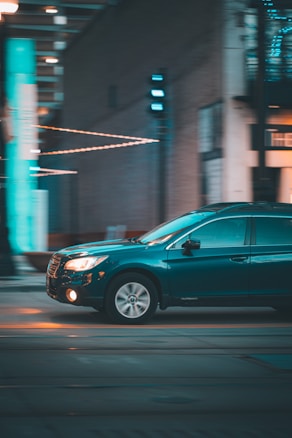 A dark teal SUV is in motion on a city street at night. The background is blurred, suggesting speed, with bright teal and orange lights reflecting off the vehicle. The road appears empty, with an urban environment featuring buildings and streetlights.