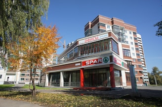 A modern multi-story building with a storefront labeled 'SPAR' in red signage dominates the foreground. The structure features large glass windows and red accents. A tall tree with autumn-colored leaves stands to the left, contrasting with the clear blue sky, while the surrounding area is lined with other buildings and pathways.