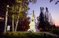 A stupa with a golden spire stands prominently amidst a garden setting surrounded by tall trees. Multicolored prayer flags are strung around it, and the scene is illuminated by a soft, warm light, suggesting it is either dawn or dusk.