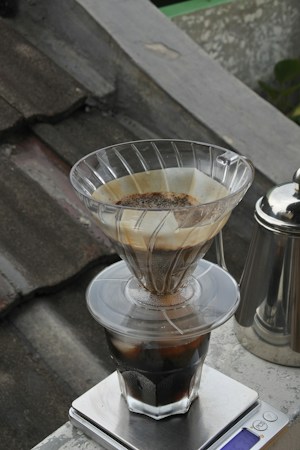 A coffee dripper situated on top of a glass container filled with ice is set atop a digital scale. The background features a weathered, tiled roof with a metal kettle visible to the side.
