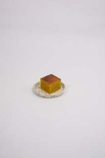 A neatly stacked, layered cake slice sits on a round, decorative plate. The cake has several visible layers of yellow and brown, giving it a textured appearance.