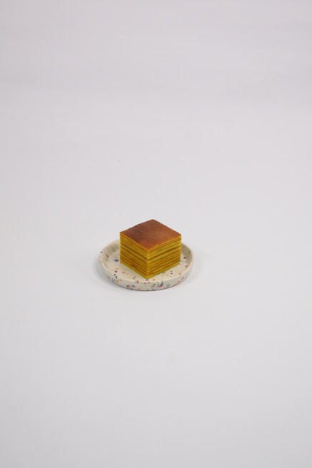 A neatly stacked, layered cake slice sits on a round, decorative plate. The cake has several visible layers of yellow and brown, giving it a textured appearance.