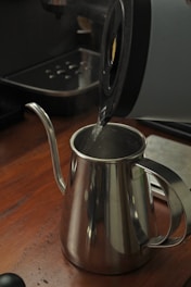 A kettle with a lid open is pouring hot water into a stainless steel gooseneck kettle on a wooden countertop. In the background, part of a coffee machine is visible, along with a tray for coffee drips.