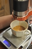 A fresh cup of espresso is being brewed, with coffee flowing from a machine into a cup positioned on a digital scale. The scale displays a weight of 28.4 grams. The machine has a copper-colored metal detailing and the cup is white with floral decorations.