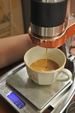 A fresh cup of espresso is being brewed, with coffee flowing from a machine into a cup positioned on a digital scale. The scale displays a weight of 28.4 grams. The machine has a copper-colored metal detailing and the cup is white with floral decorations.