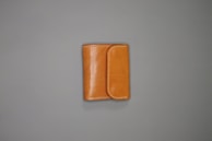 A tan leather wallet is placed on a plain gray surface. The wallet features white stitching along the edges and has a flap closure.