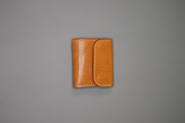 A tan leather wallet is placed on a plain gray surface. The wallet features white stitching along the edges and has a flap closure.