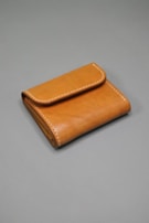 A brown leather wallet with visible stitching along the edges is placed on a plain gray surface. The wallet is neatly folded, showcasing its compact and minimalist design.