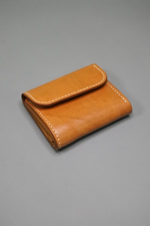 A brown leather wallet with visible stitching along the edges is placed on a plain gray surface. The wallet is neatly folded, showcasing its compact and minimalist design.