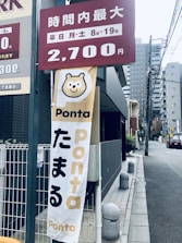 A vertical banner with a cartoon bear face and the word 'Ponta' displayed prominently. Below the bear face, more text is visible. Above, there is a maroon sign indicating pricing information in Japanese. The scene is set on a city street with tall buildings and electrical wires in the background.